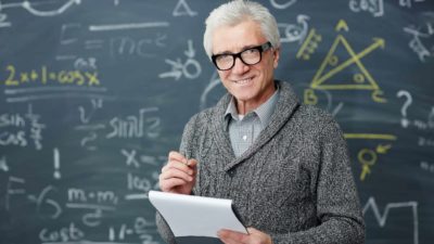 A grey-haired mature-aged man with glasses stands in front of a blackboard filled with mathematical workings as he holds a pad of paper in one hand and a pen in the other and stands smiling at the camera.